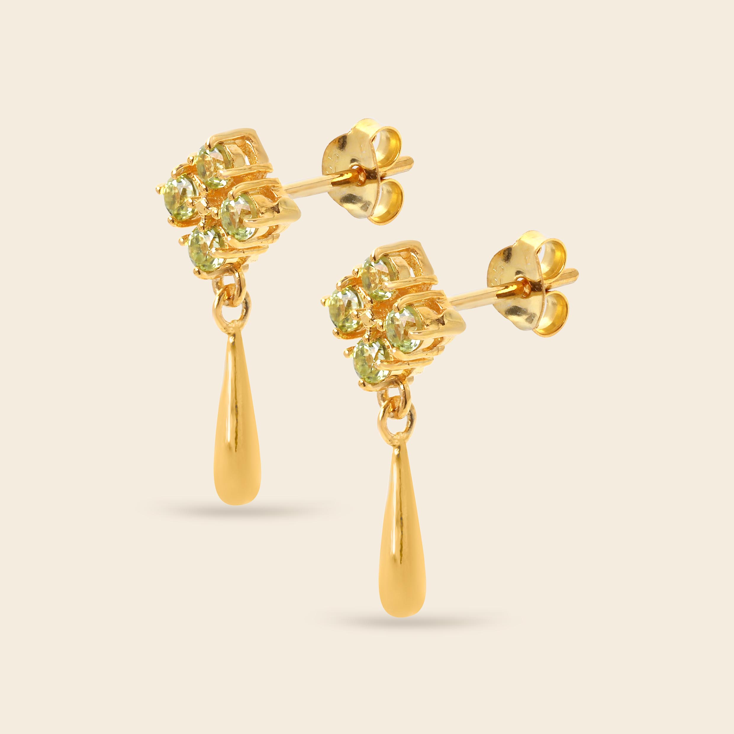 A pair of gold dangle earrings featuring a cluster of round-cut light green gemstones in a floral setting, with a polished gold teardrop drop hanging below, shown on a light beige background.