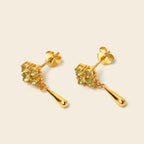 A pair of gold drop earrings with a cluster of small green gemstones and a teardrop-shaped drop, shown on a light beige background.