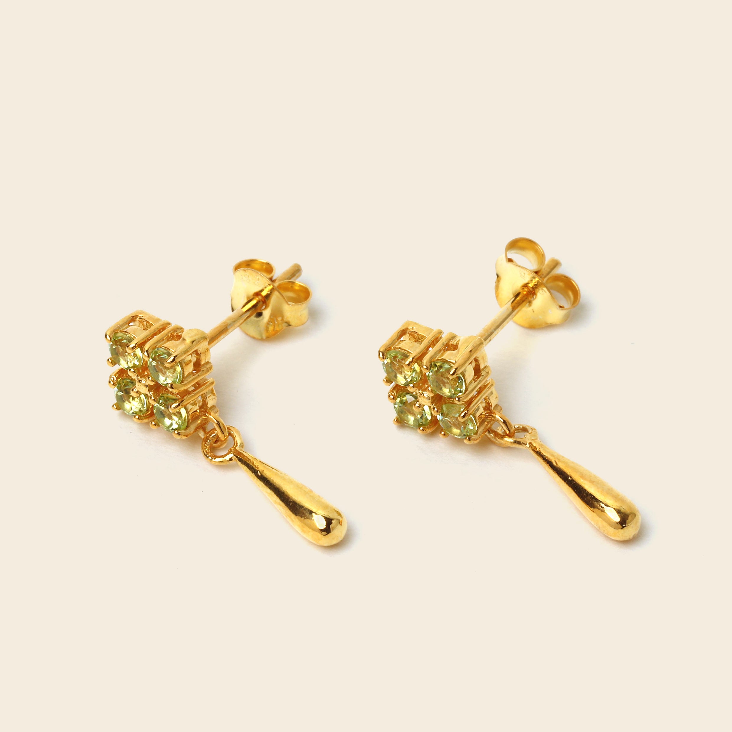 A pair of gold drop earrings with a cluster of small green gemstones and a teardrop-shaped drop, shown on a light beige background.