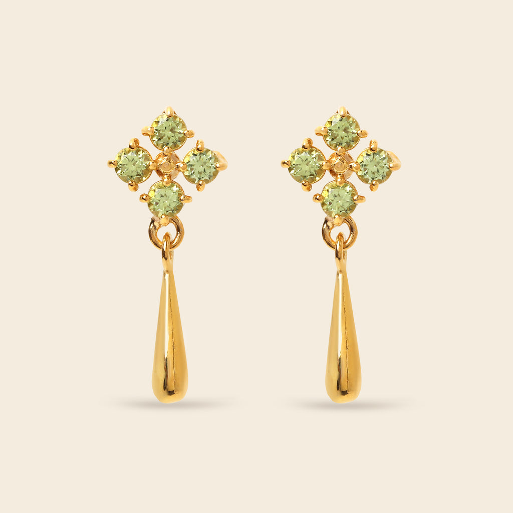 A pair of gold dangle earrings featuring a four-stone floral cluster in light green peridot-like gemstones above a polished gold teardrop drop, set against a solid light beige background.
