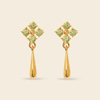 A pair of gold dangle earrings featuring a four-stone floral cluster in light green peridot-like gemstones above a polished gold teardrop drop, set against a solid light beige background.