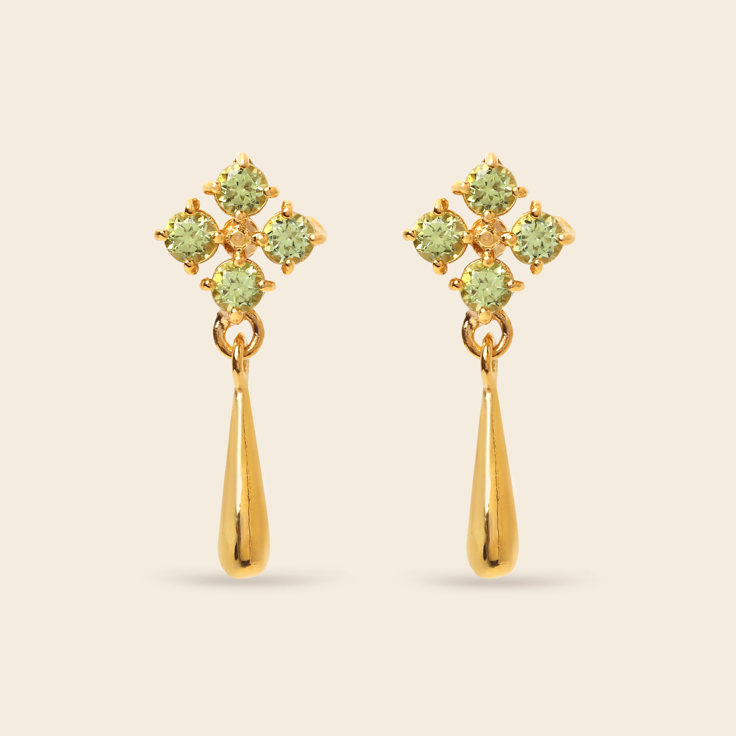 A pair of gold dangle earrings featuring a four-stone floral cluster in light green peridot-like gemstones above a polished gold teardrop drop, set against a solid light beige background.