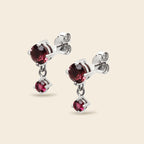 A pair of silver stud earrings with heart-shaped dark red gemstones and a smaller dangling heart gemstone, shown on a light beige background.