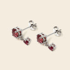 A pair of silver stud earrings with a large round red gemstone and a smaller dangling red gemstone, set against a solid light beige background.