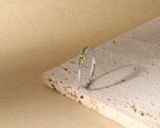 A slim silver band ring with a row of small green gemstones, displayed on a textured beige stone slab against a warm beige background.