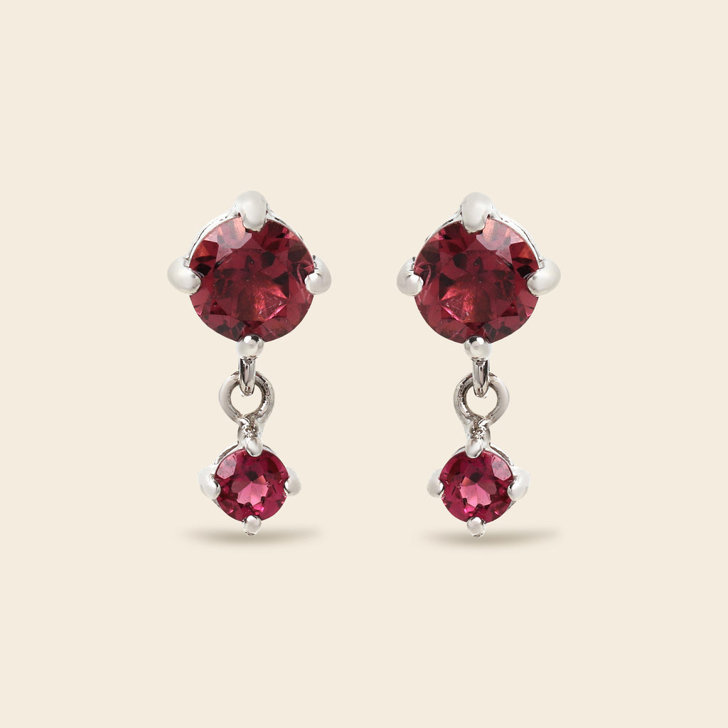 A pair of silver drop earrings featuring large round-cut deep red gemstones on the top and smaller oval-cut red gemstones below, set against a solid light beige background.