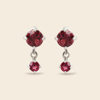 A pair of silver drop earrings featuring large round-cut deep red gemstones on the top and smaller oval-cut red gemstones below, set against a solid light beige background.