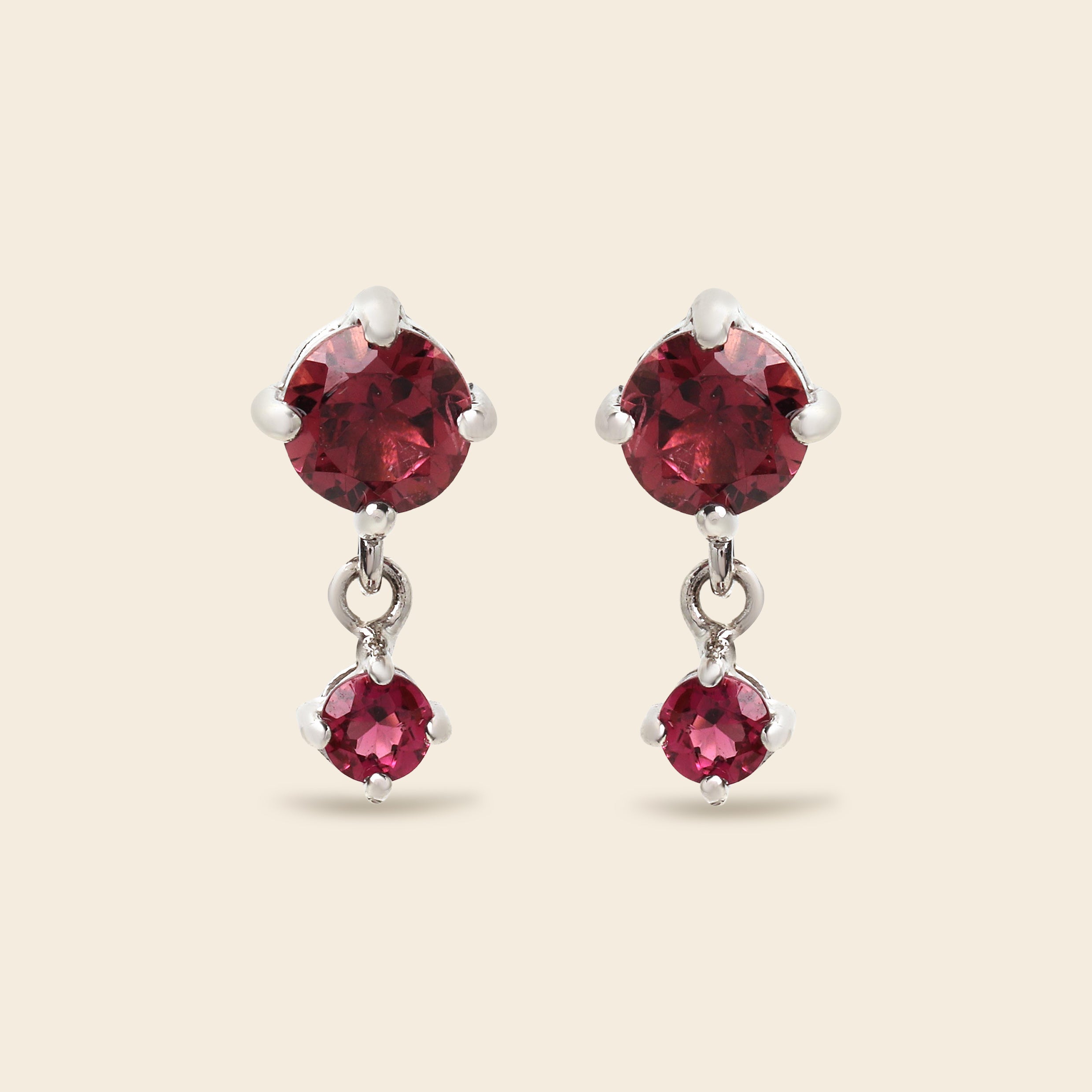 A pair of silver drop earrings featuring large round-cut deep red gemstones on the top and smaller oval-cut red gemstones below, set against a solid light beige background.