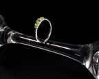 A silver ring with a small cluster of green gemstones, displayed on a clear glass stem against a black background.