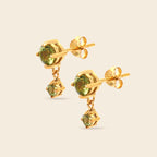 A pair of gold stud earrings with round green gemstones and smaller dangling green gemstone charms, shown on a light beige background.