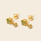 A pair of gold stud earrings with round light green gemstones and a smaller dangling green accent stone, shown on a plain beige background.