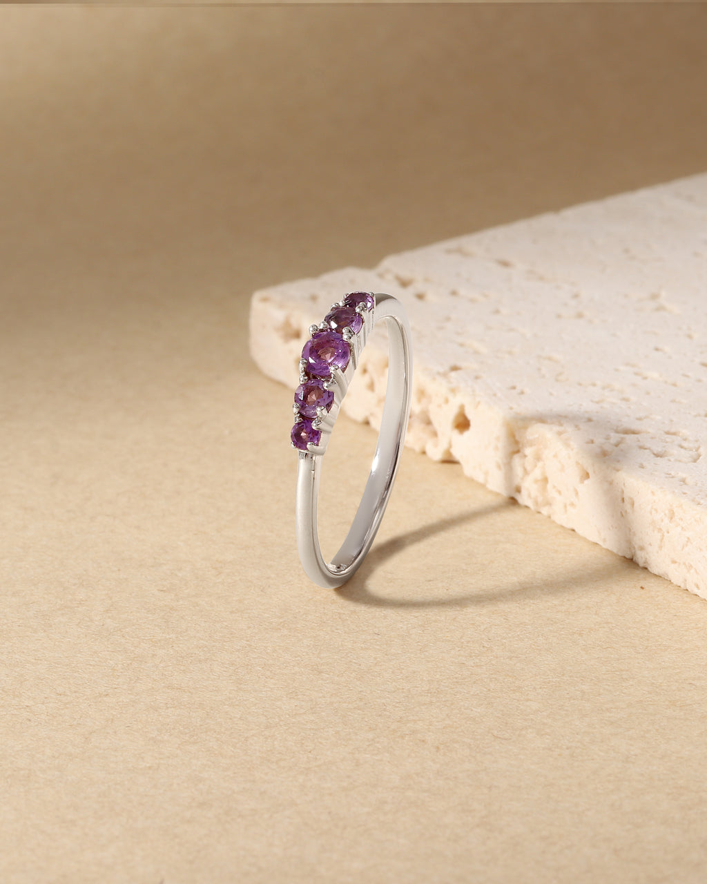 A silver band ring featuring a row of small purple gemstones, displayed on a textured beige surface next to a light stone prop.
