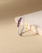 A silver band ring featuring a row of small purple gemstones, displayed on a textured beige surface next to a light stone prop.