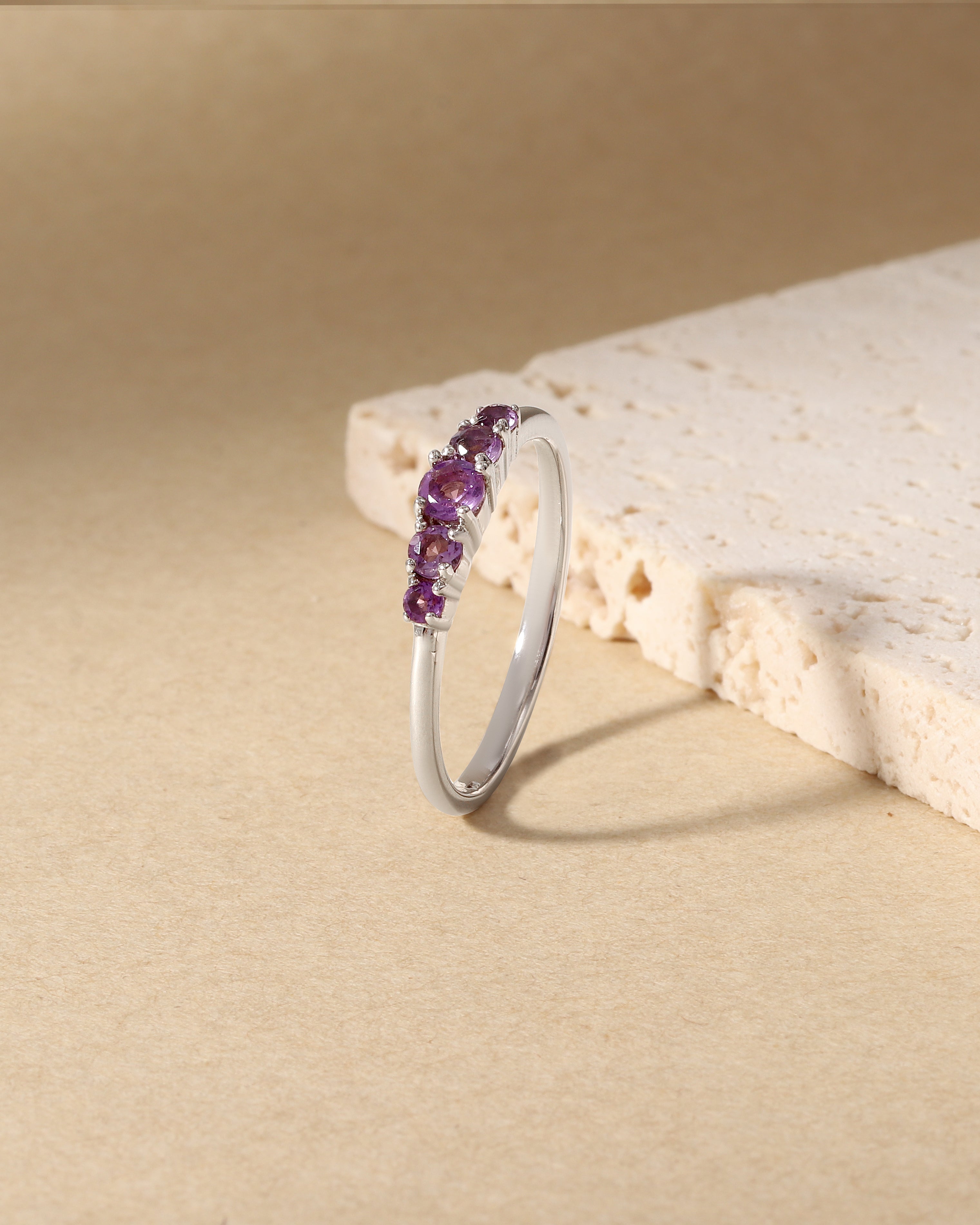 A silver band ring featuring a row of small purple gemstones, displayed on a textured beige surface next to a light stone prop.