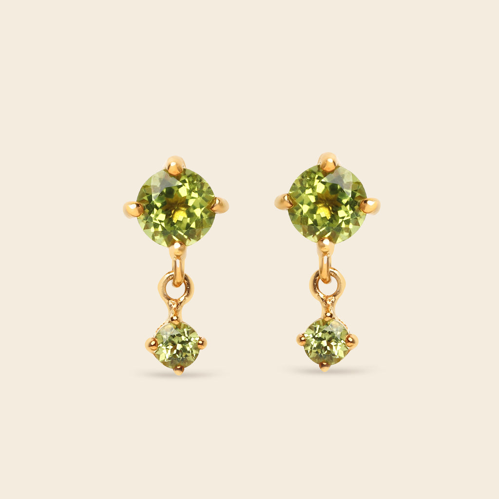 A pair of gold drop earrings featuring round green peridot gemstones, with a larger round stone on the top and a smaller prong-set stone dangling below, set against a solid light beige background.