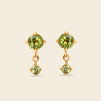 A pair of gold drop earrings featuring round green peridot gemstones, with a larger round stone on the top and a smaller prong-set stone dangling below, set against a solid light beige background.