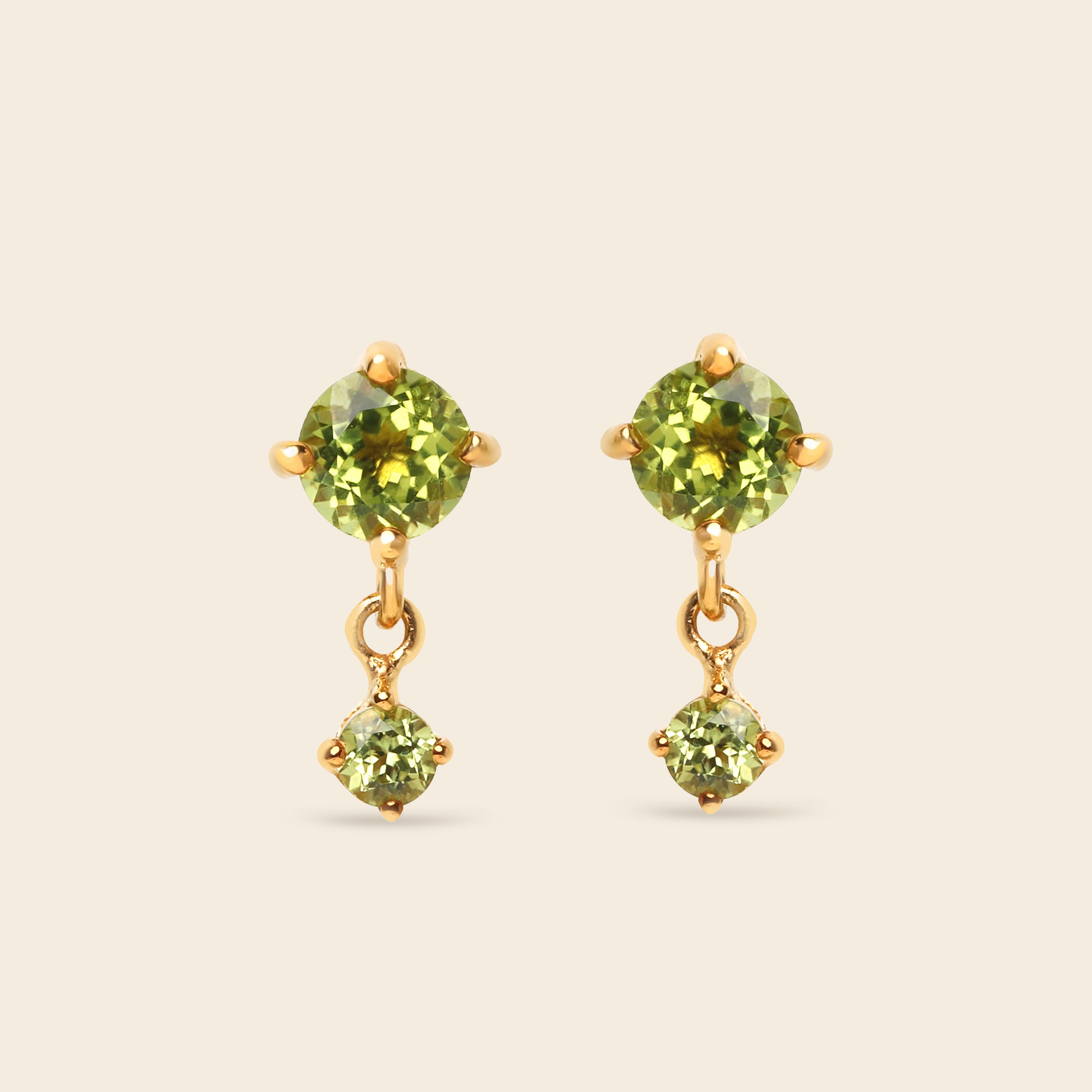 A pair of gold drop earrings featuring round green peridot gemstones, with a larger round stone on the top and a smaller prong-set stone dangling below, set against a solid light beige background.