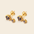A pair of gold stud earrings with oval blue gemstones and a small square blue accent stone, shown on a light beige background.