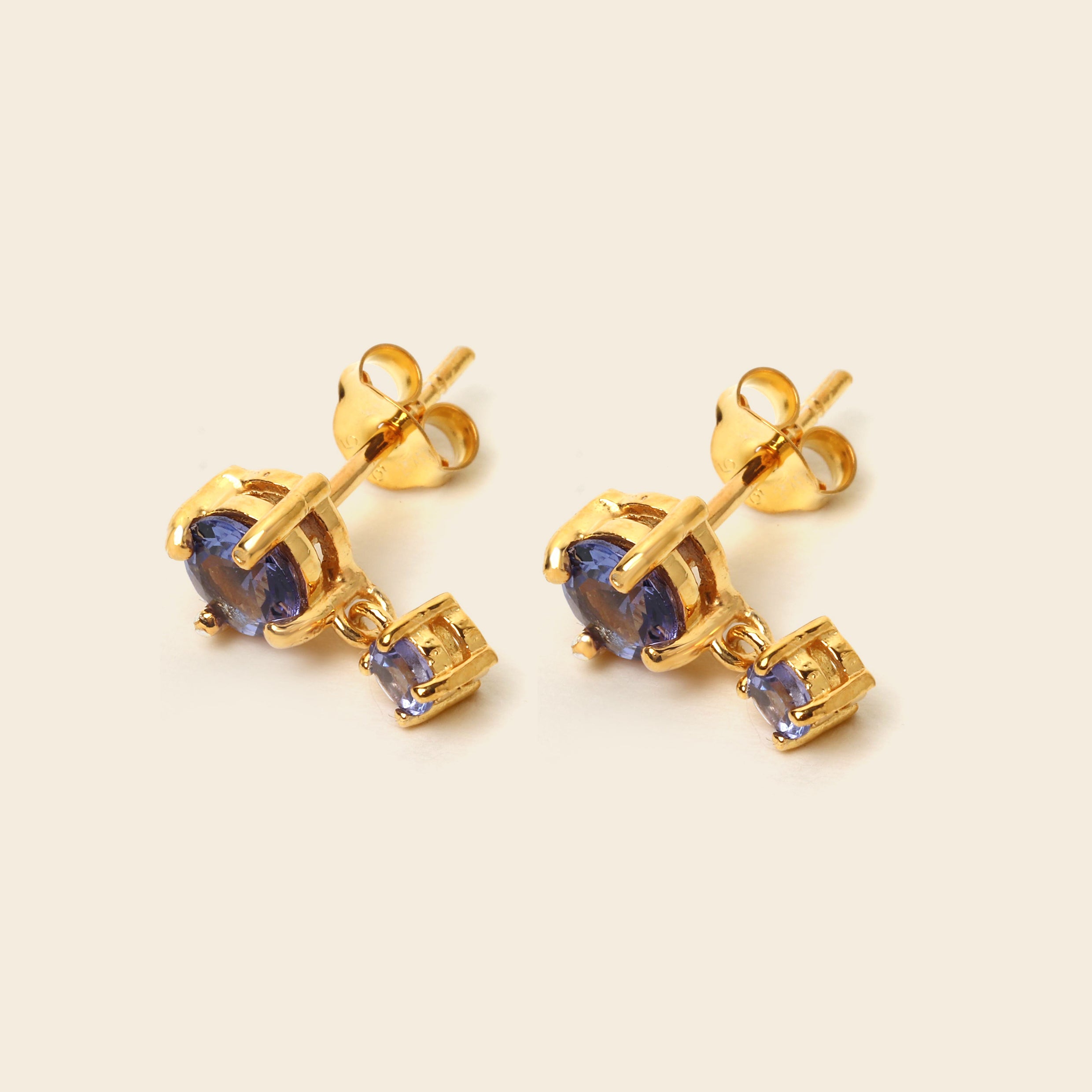 A pair of gold stud earrings with oval blue gemstones and a small square blue accent stone, shown on a light beige background.