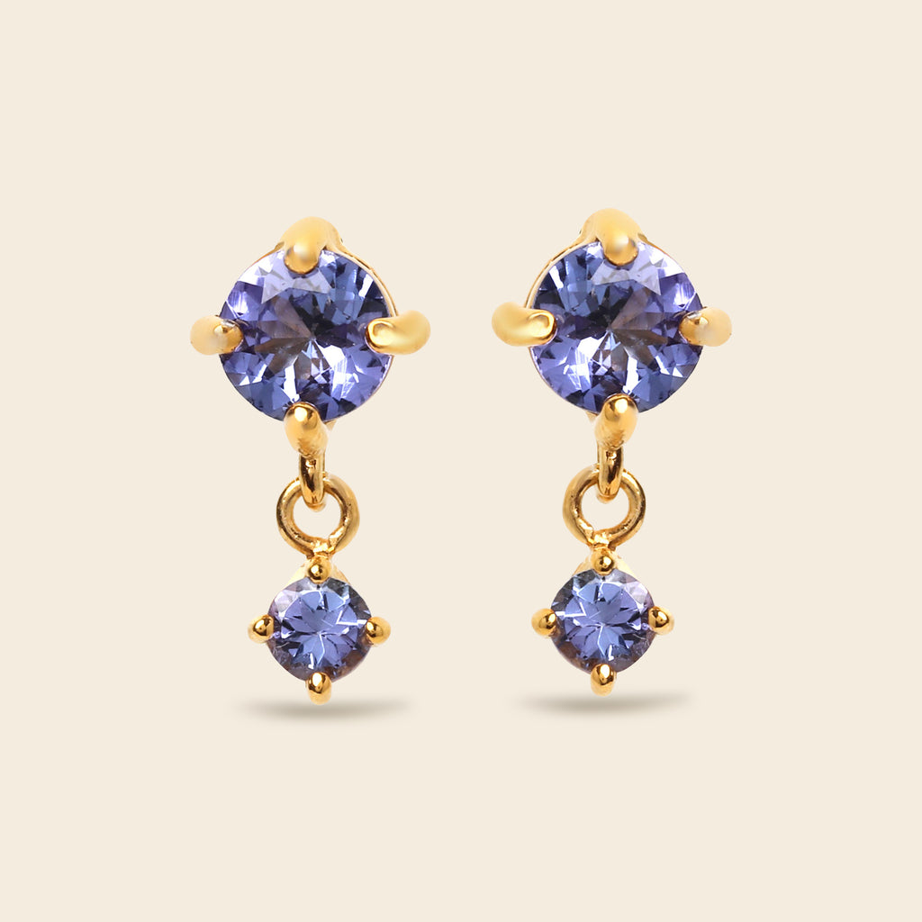 A pair of gold drop earrings featuring round-cut blue-purple gemstones, shown on a solid light beige background.