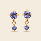 A pair of gold drop earrings featuring round-cut blue-purple gemstones, shown on a solid light beige background.