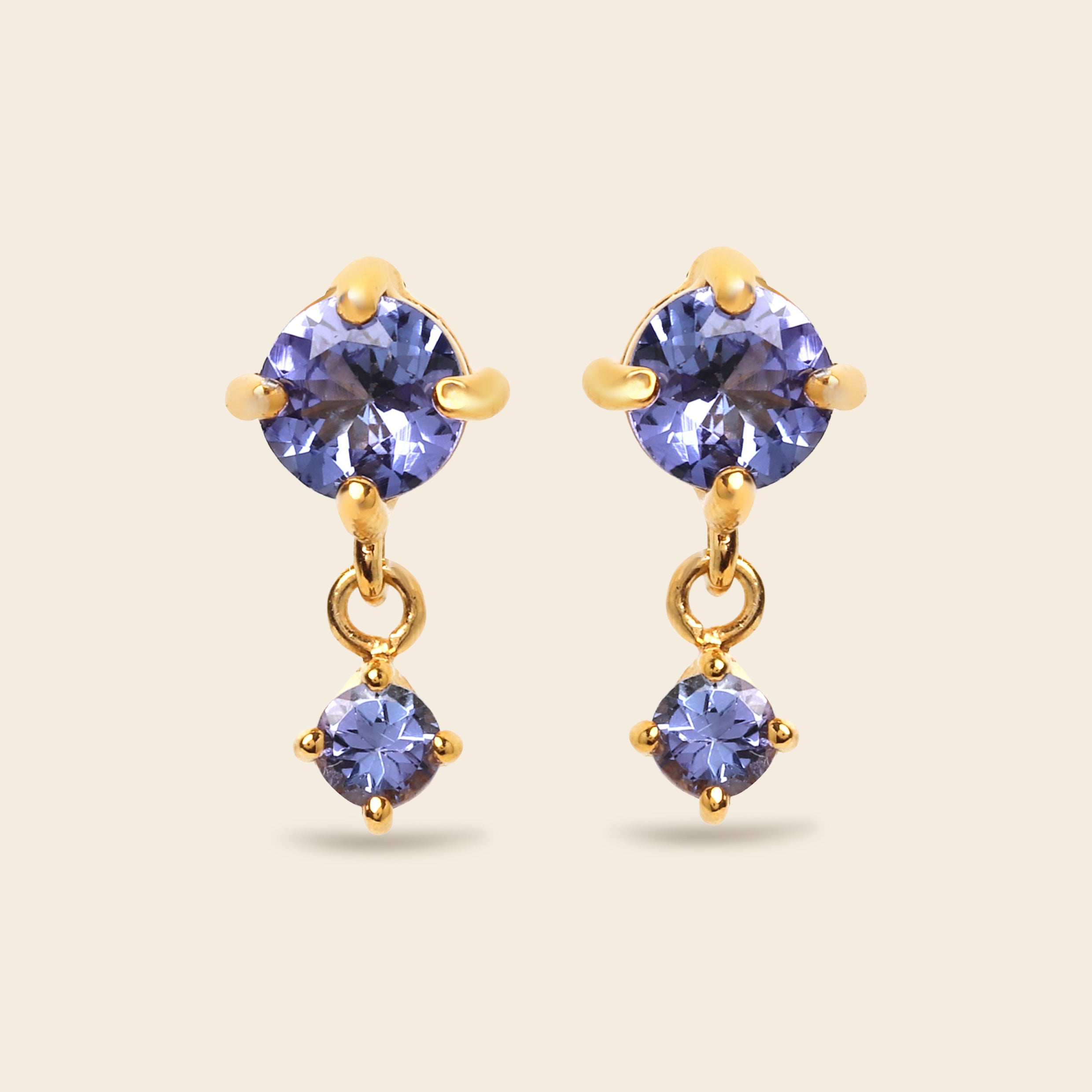 A pair of gold drop earrings featuring round-cut blue-purple gemstones, shown on a solid light beige background.