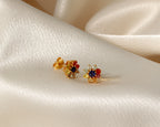 A pair of gold-toned stud earrings with colorful gemstone clusters, resting on draped cream satin fabric.