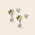 A pair of silver stud earrings with oval green gemstones and smaller dangling round green gemstones, shown on a light beige background.