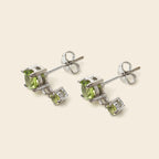 A pair of silver stud earrings featuring a large round green peridot gemstone and a smaller rectangular green gemstone, set in silver-toned metal on a light beige background.