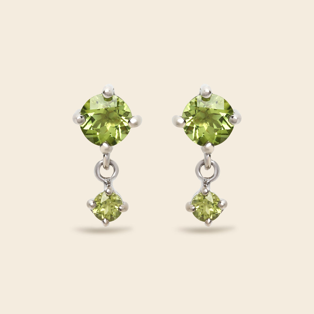 A pair of silver drop earrings featuring round green peridot gemstones, shown against a solid light beige background.