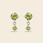 A pair of silver drop earrings featuring round green peridot gemstones, shown against a solid light beige background.