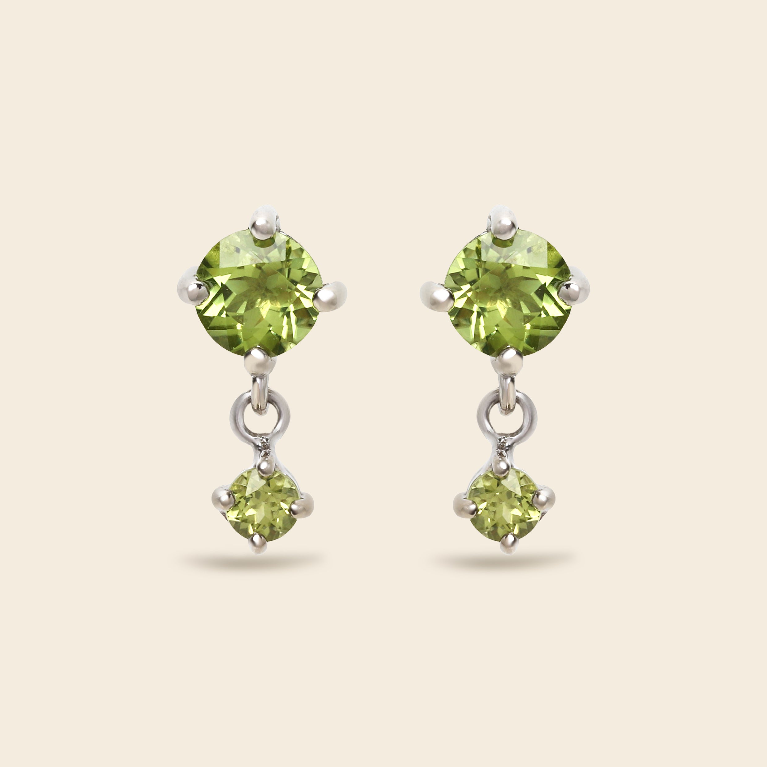 A pair of silver drop earrings featuring round green peridot gemstones, shown against a solid light beige background.