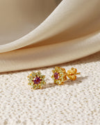 A pair of gold flower-shaped stud earrings with small clear stones and a central purple gemstone, displayed on a textured cream surface with draped satin fabric in the background.