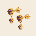 A pair of gold drop stud earrings with round purple gemstones, shown on a light beige background.
