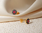 A pair of gold floral stud earrings with a central pink gemstone surrounded by dark blue stones, displayed on draped cream satin fabric and a textured cream surface.