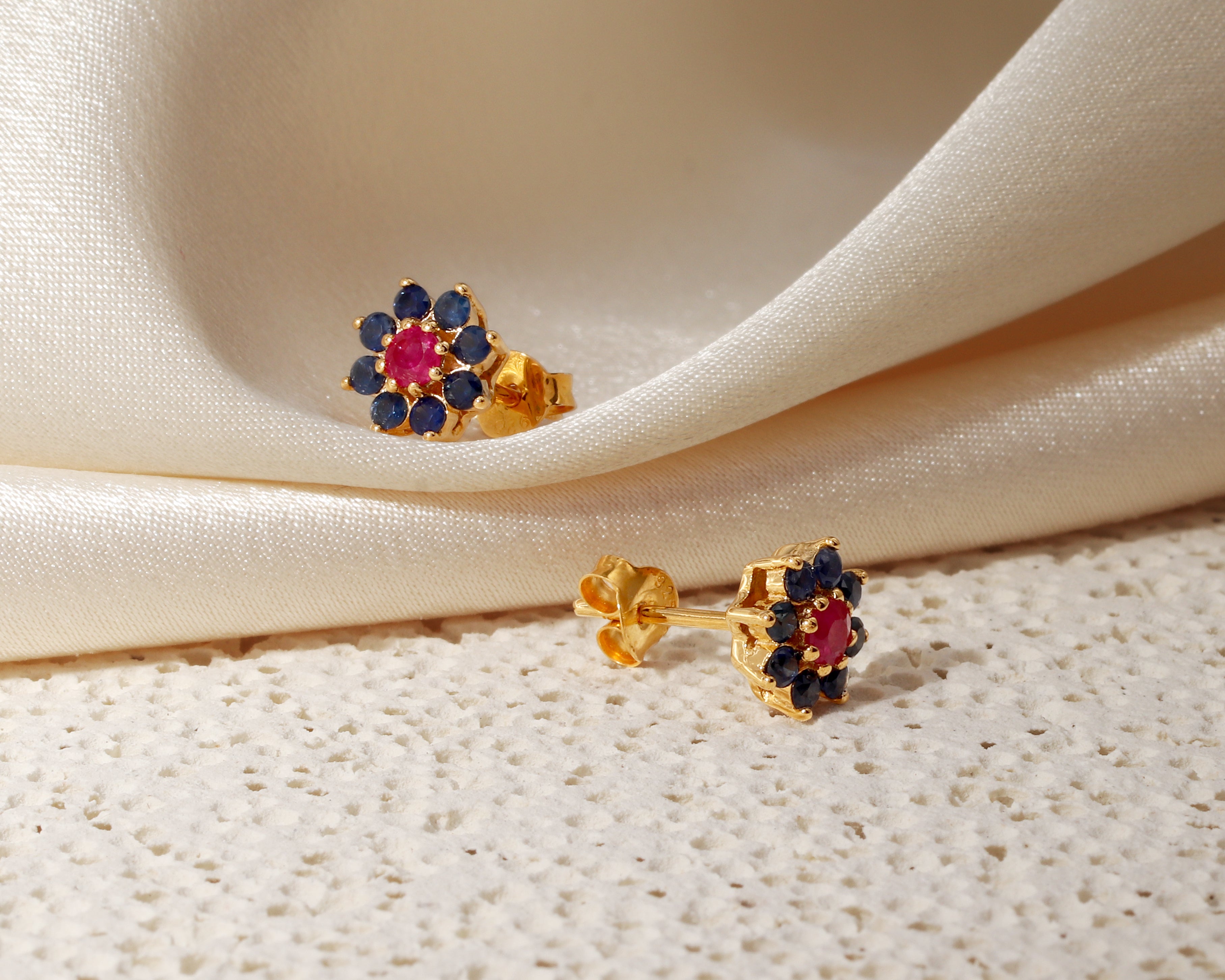 A pair of gold floral stud earrings with a central pink gemstone surrounded by dark blue stones, displayed on draped cream satin fabric and a textured cream surface.