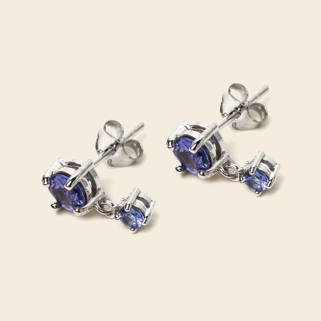 A pair of silver stud earrings with a large oval blue gemstone and a smaller round blue gemstone drop, shown on a light beige background.