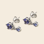 A pair of silver stud earrings with a large oval blue gemstone and a smaller round blue gemstone drop, shown on a light beige background.