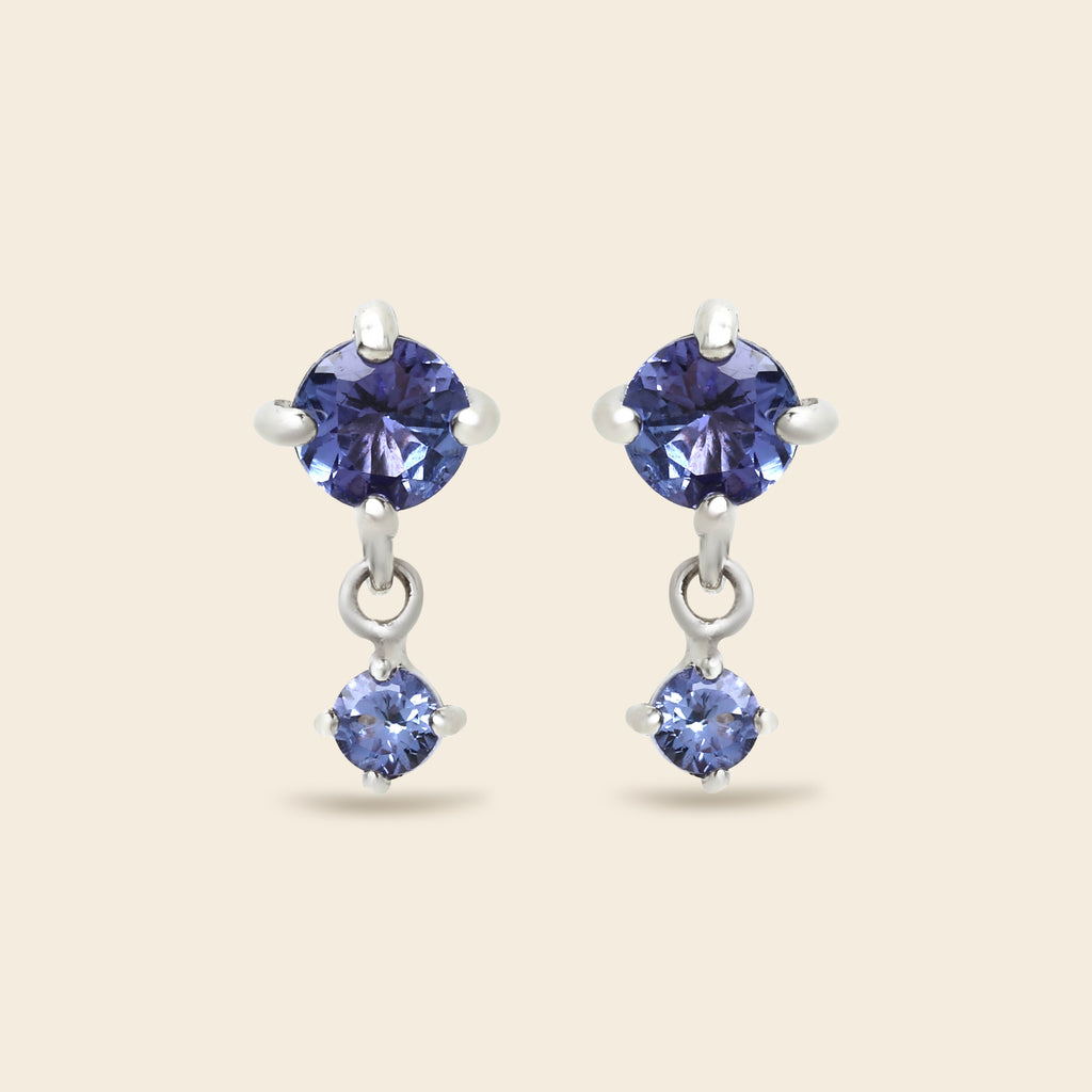 A pair of silver drop earrings featuring round-cut blue gemstones, shown against a solid light beige background.