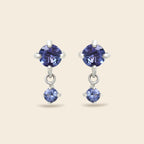 A pair of silver drop earrings featuring round-cut blue gemstones, shown against a solid light beige background.
