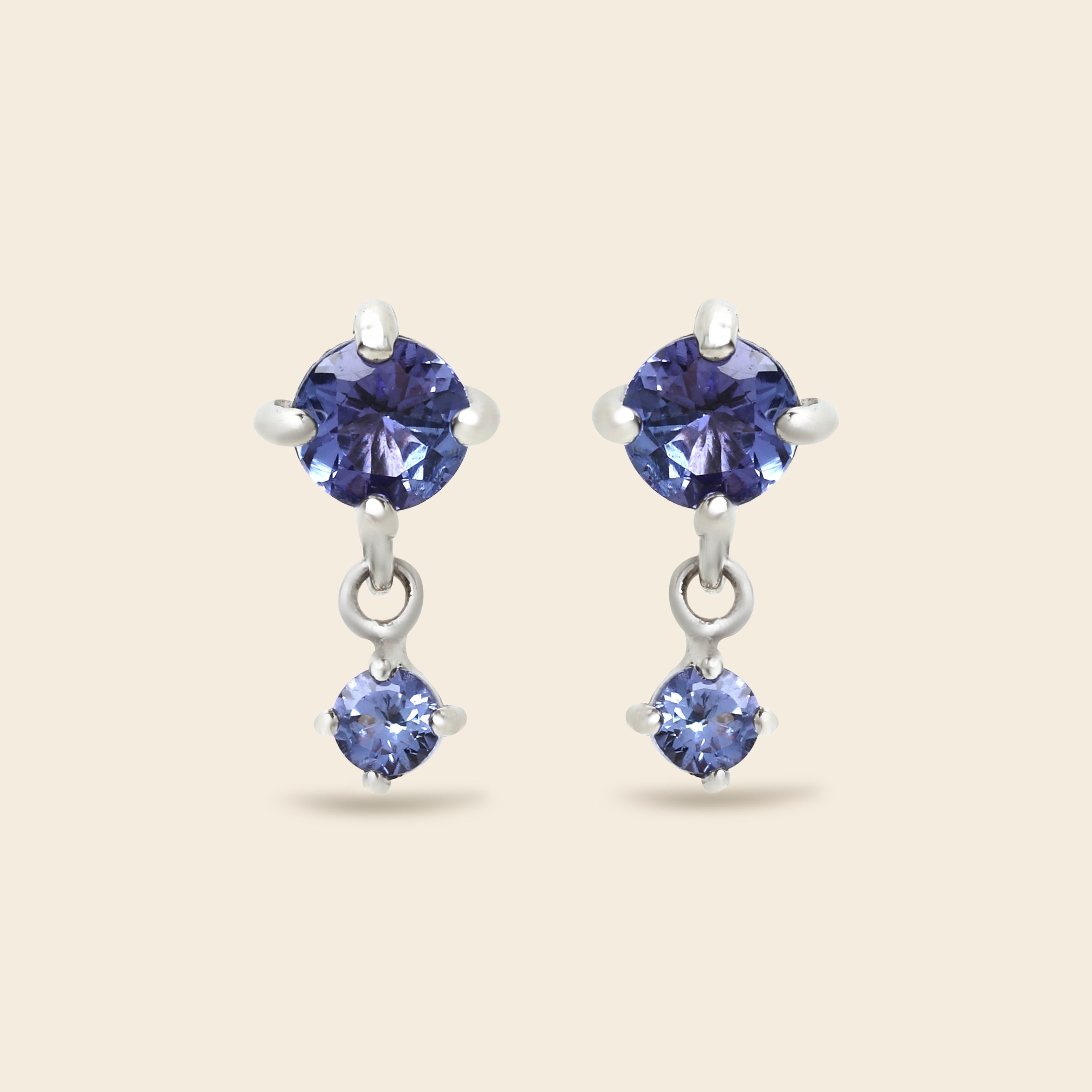 A pair of silver drop earrings featuring round-cut blue gemstones, shown against a solid light beige background.