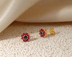 A pair of gold flower-shaped stud earrings with a dark blue center and surrounding red gemstone halo, displayed on a textured beige surface with a draped cream satin fabric in the background.
