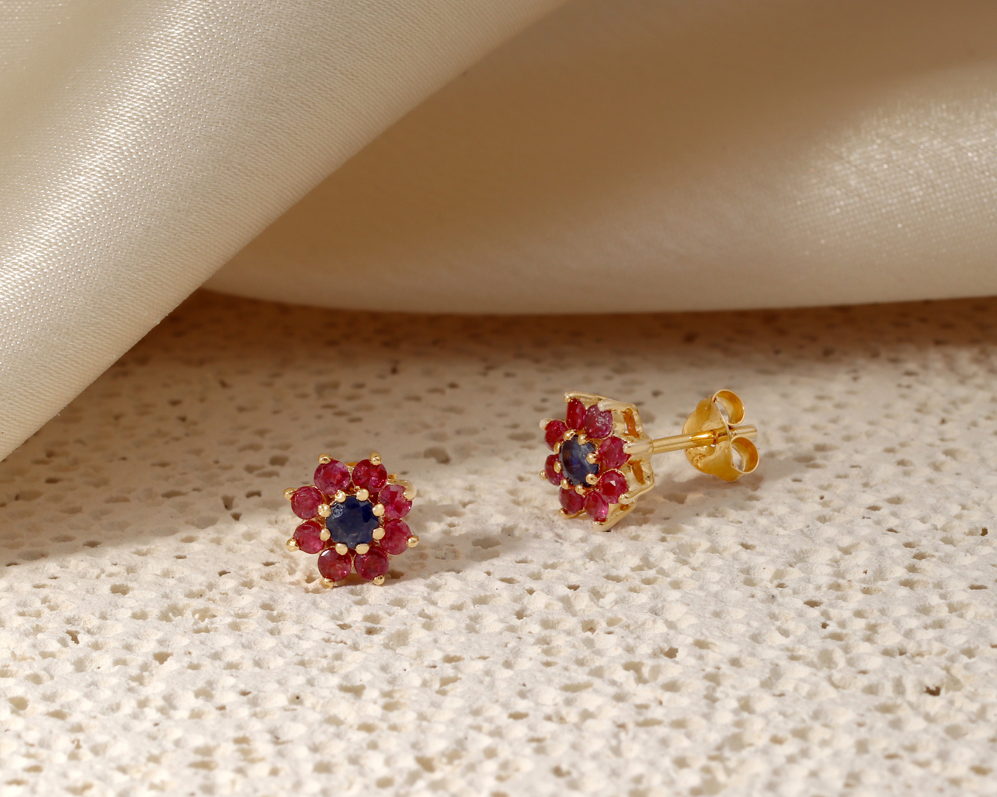 A pair of gold flower-shaped stud earrings with a dark blue center and surrounding red gemstone halo, displayed on a textured beige surface with a draped cream satin fabric in the background.