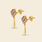 A pair of gold dangle earrings with a small cluster of light purple gemstones at the top and a teardrop-shaped gold drop below, shown on a light beige background.