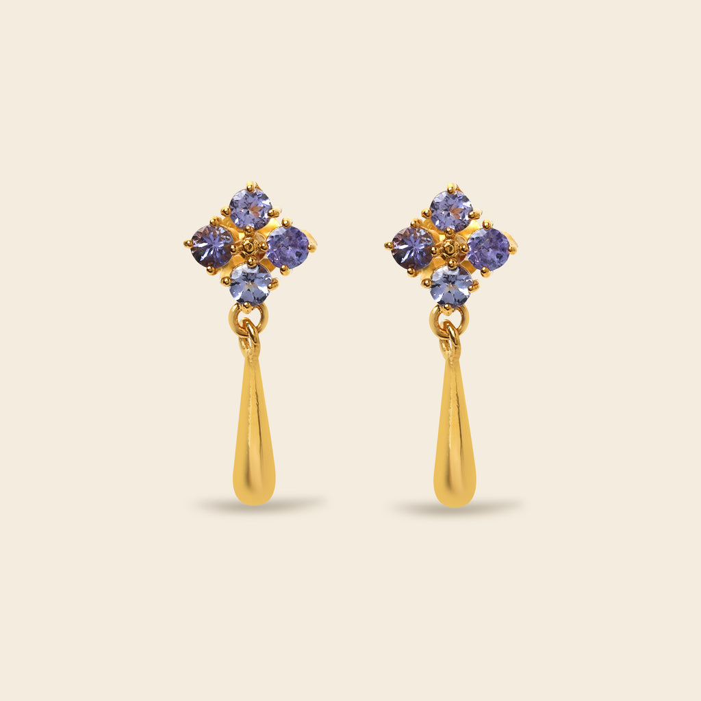 A pair of gold dangle earrings featuring a cluster of five round-cut purple gemstones at the top and a polished gold teardrop-shaped drop below, set against a solid light beige background.