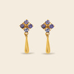 A pair of gold dangle earrings featuring a cluster of five round-cut purple gemstones at the top and a polished gold teardrop-shaped drop below, set against a solid light beige background.