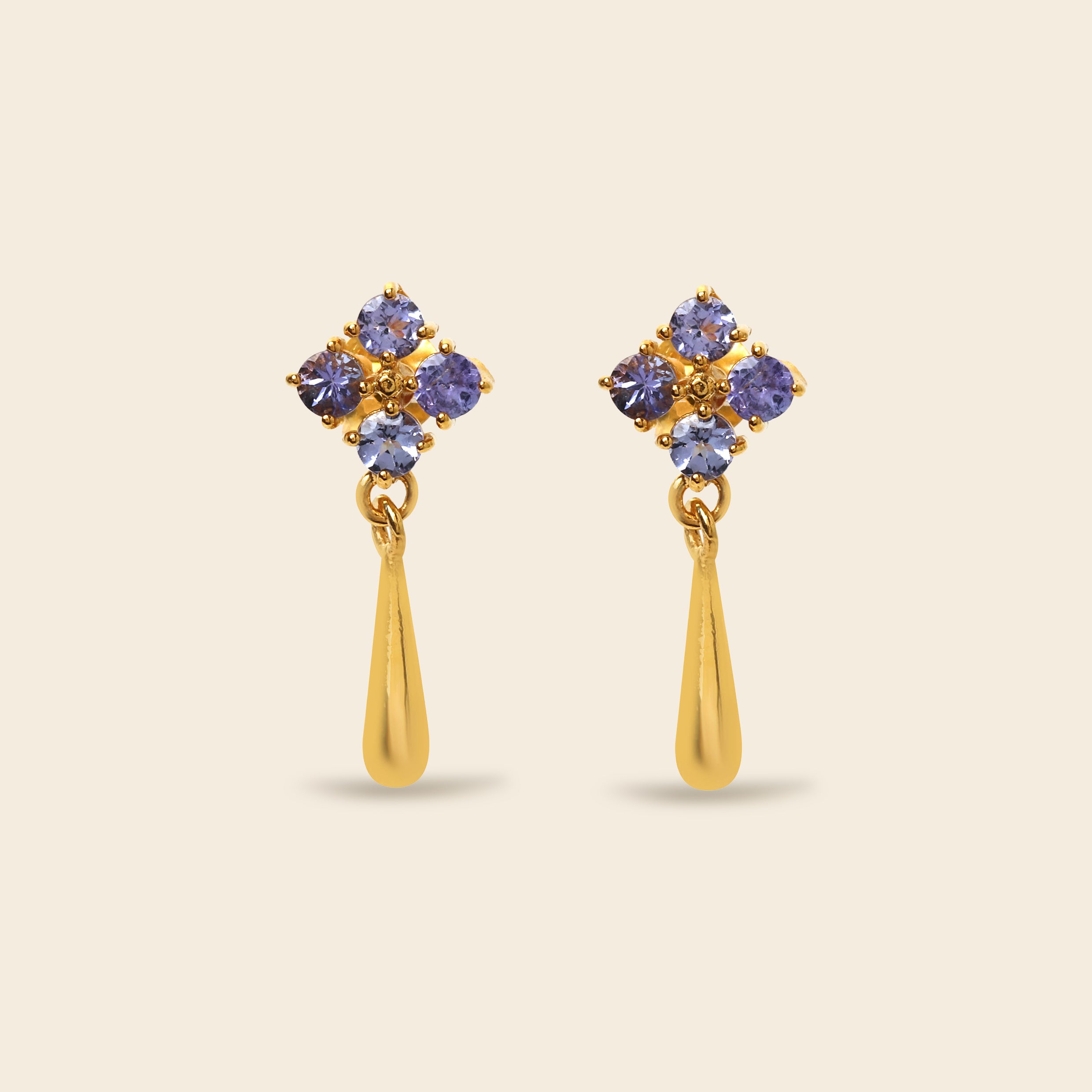 A pair of gold dangle earrings featuring a cluster of five round-cut purple gemstones at the top and a polished gold teardrop-shaped drop below, set against a solid light beige background.