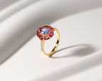 A gold ring featuring a round blue center stone surrounded by a halo of pink gemstones, with small clear stones along the band, set against a white draped background.