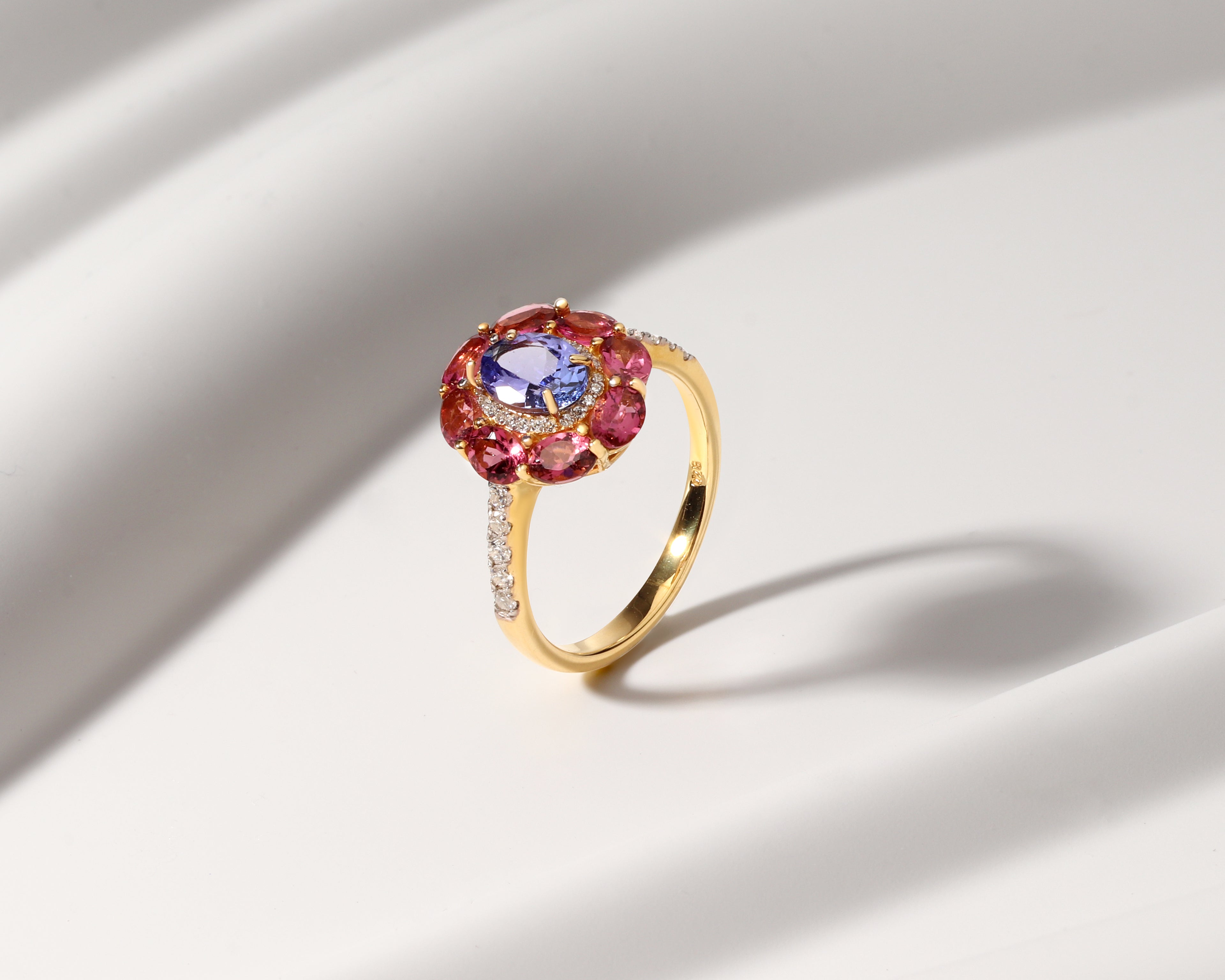 A gold ring featuring a round blue center stone surrounded by a halo of pink gemstones, with small clear stones along the band, set against a white draped background.