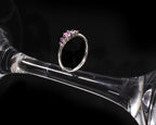 A silver ring with a row of small pink gemstones, displayed on the stem of a clear glass goblet against a black background.